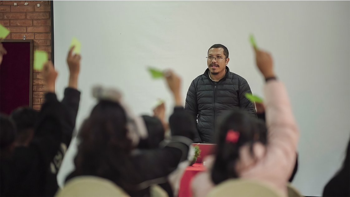 A photo of a man standing at the front of a room. In the foreground are people raising their hand and holding green paper.