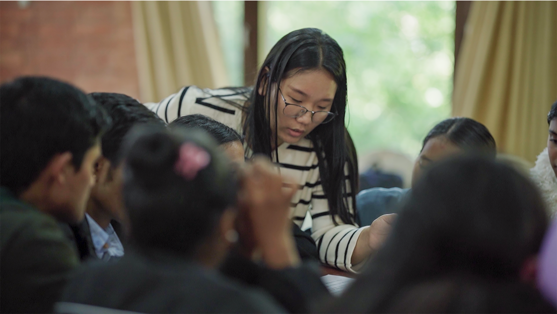 A photo of a woman standing by a group of children sat at a table.