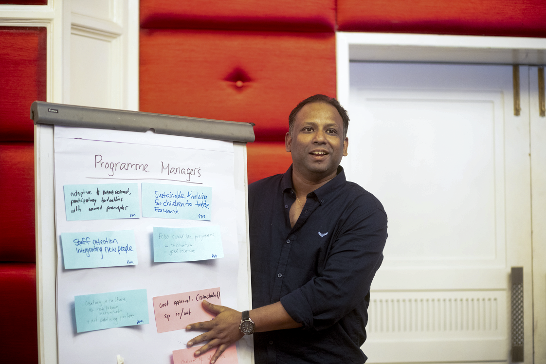 A photo of a man standing next to a flipboard with notes from research meeeting, speaking to the wider room.