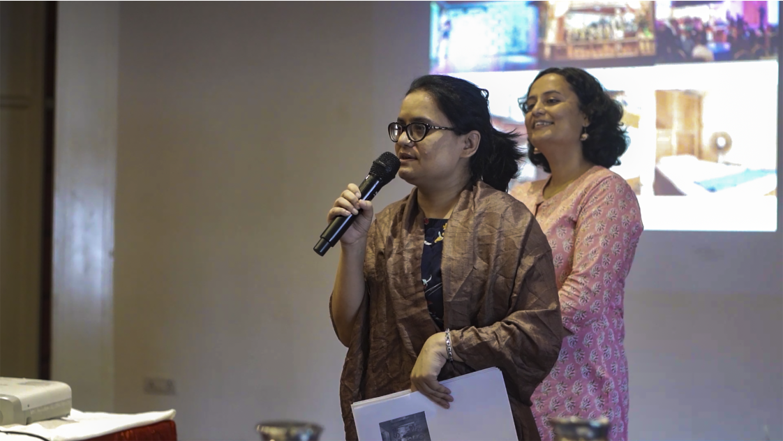 A photo of two women speaking to a crowd, standing in front of a presentation and holding a microphone.