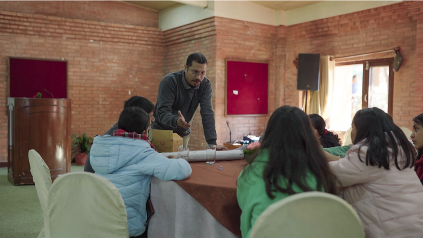 A photo of several girls sat around a table with an adult man speaking to them, engaged in conversation