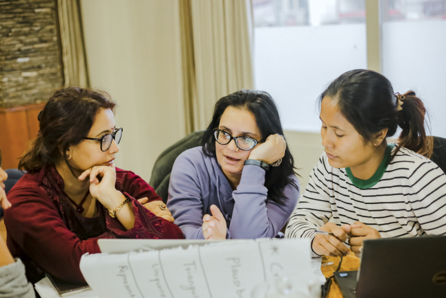 A photo of three women sat at a desk engaged in conversation.