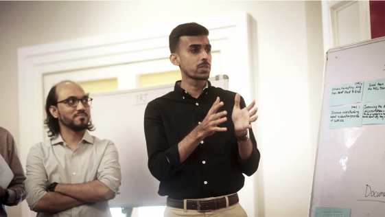 A photo of two men standing in front of a whiteboard, speaking to a room of people.