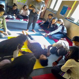 A photo taken in a workshop. Children are sat around the outside with one in the middle, doing a body mapping exercise.