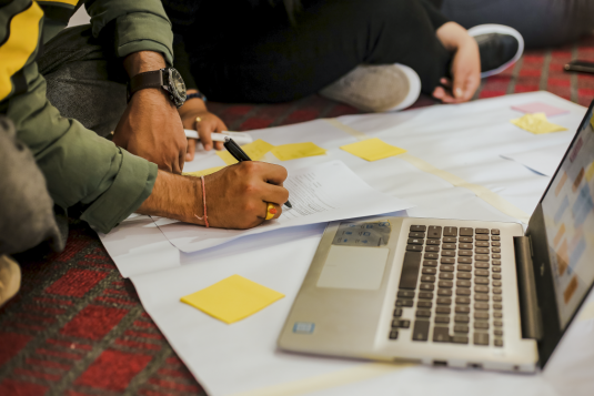 A photo of a mans hand writing on a piece of paper with a laptop by his side.